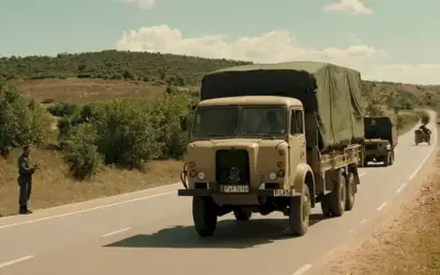 Camión que transporta mercancías en la carretera abierta con personal militar y suministros en segundo plano. Texto español al inglés: ¿Cómo satisfacen las necesidades de suministro durante la guerra? (Frativa española